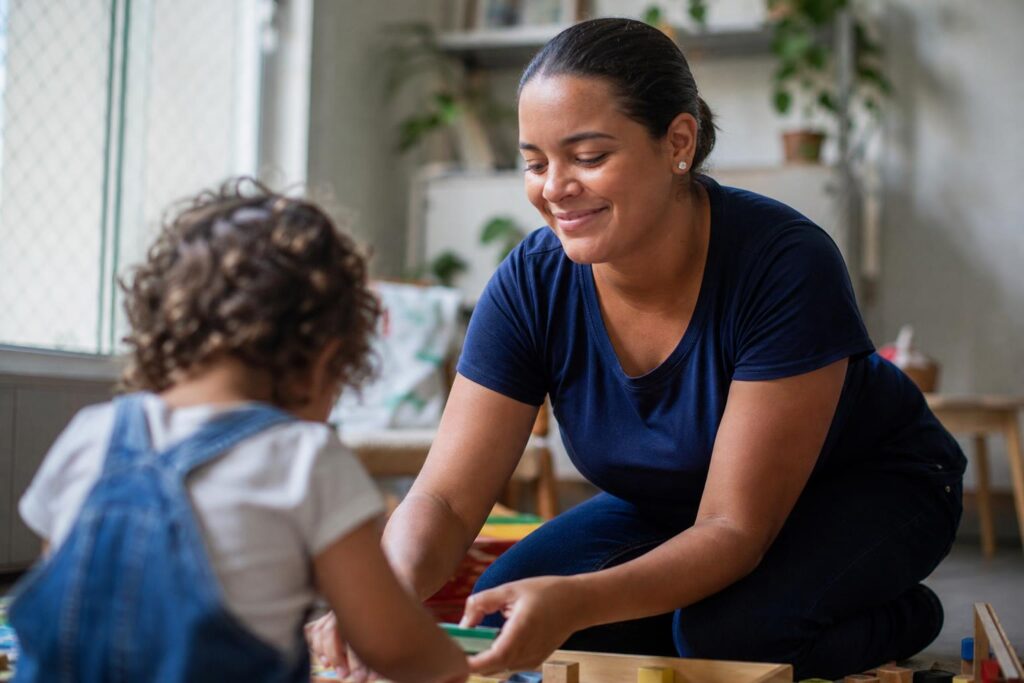 Babá sorridente ajoelhada no chão brincando com uma criança pequena com blocos de madeira em um quarto infantil iluminado.