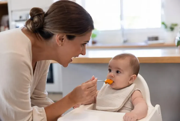 Mulher sorridente oferecendo uma colher com purê de cenoura para um bebê de 6 meses sentado em um cadeirão de alimentação com babador.