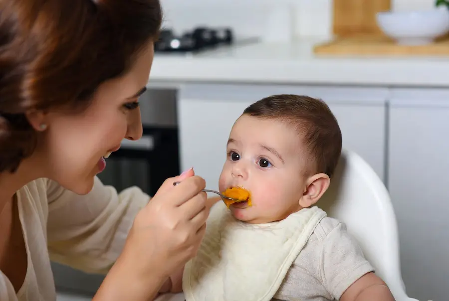 Mulher sorridente com cabelos presos segura uma colher e oferece um purê de cenoura para um bebê de 6 meses sentado em um cadeirão de alimentação branco, com babador e olhando para a colher.