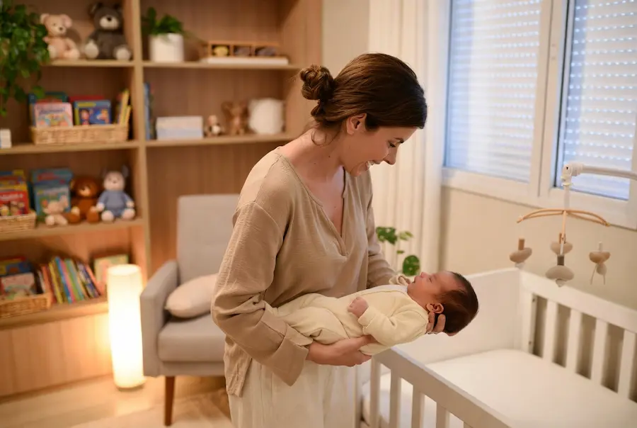 rotina-de-sono-para-bebe-de-6-meses Mãe brasileira de pele morena clara, cabelos castanhos presos em coque baixo, em pé no quarto infantil à noite, segurando com delicadeza seu bebê de cerca de 6 meses já adormecido, vestido com macacão bege, enquanto o prepara para deitar no berço branco. Iluminação quente e suave, ambiente aconchegante com estante de livros, pelúcias e móbile ao fundo.