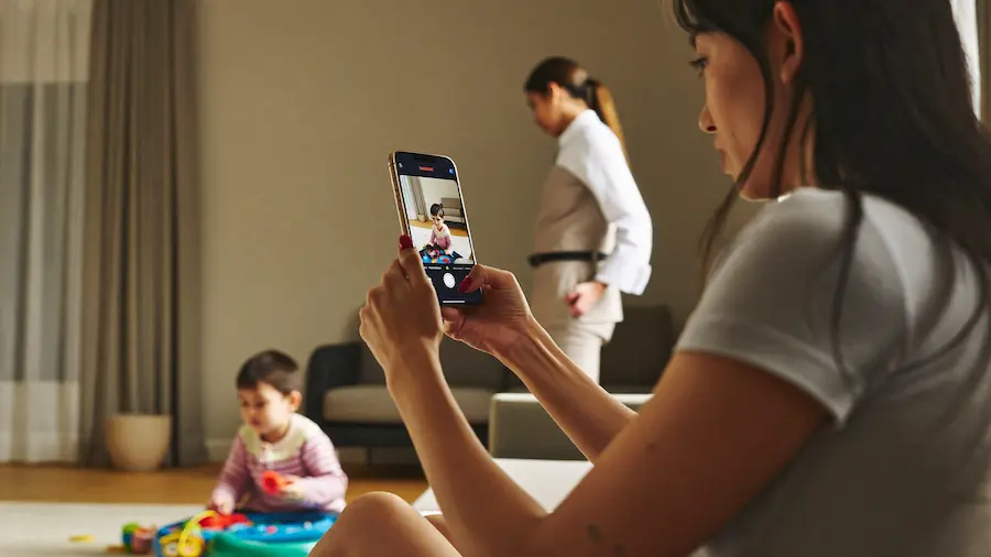 mãe verificando câmera de babá pelo celular enquanto criança brinca sozinha em casa